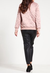Chaqueta bomber acolchada rosa con ribete acanalado, combinada con pantalones de piel sintética negra y zapatos de tacón alto negros. Vista trasera.