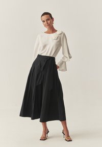 Biała bluzka z długim rękawem z kwiatowym akcentem; czarna spódnica do talii z plisami; czarne sandały na obcasie z paskami; minimalistyczny styl na jasnym tle.