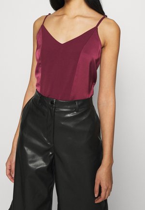 Vrouw met een mouwloze satijnen top in bordeauxrood met dunne bandjes, gestopt in een zwarte leren broek met hoge taille.