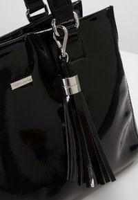 Sac à main noir brillant avec un pompon en cuir amovible et une quincaillerie argentée portant la marque "Tamaris" sur le sac et le pompon.