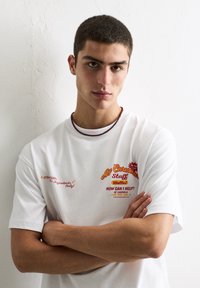 Hvid bomulds-T-shirt med fed, farverig tekst; designet inkluderer et kok-logo og flere sætninger i rød og gul. Glat tekstur.