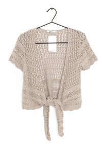 Beige kortärmad virkade kofta med knytframsida, hängande på svart galge mot vit bakgrund.