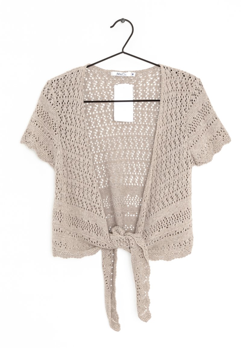 Beige kortärmad virkade kofta med knytframsida, hängande på svart galge mot vit bakgrund.