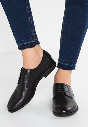 Chaussures oxford en cuir noir avec une texture lisse, bout arrondi et détails de laçage, portées avec un jean en denim foncé à ourlet effiloché.