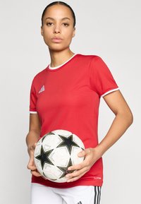 Κόκκινο αθλητικό πουκάμισο με λευκή περιφέρεια στο γιακά και λογότυπο με τρεις ρίγες, σε συνδυασμό με ποδοσφαιρική μπάλα μαύρου και λευκού χρώματος με αστέρια, κρατημένη στα χέρια.