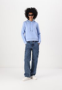 Ανοιχτού μπλε φούτερ με μπροστινή τσέπη και κορδόνια, συνδυασμένο με φαρδιά μπλέ cargo τζιν με πλαϊνές τσέπες και λευκά αθλητικά παπούτσια.