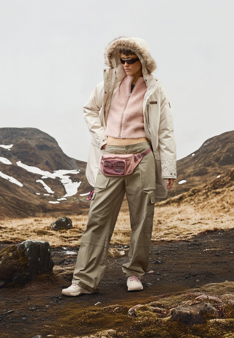 Beiger Parka mit Fellbesatz an der Kapuze, pinkem Pullover, beigen Cargo-Hosen und hellrosa Bauchtasche. Das Model trägt Sonnenbrille und neutrale Schuhe.