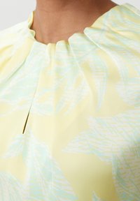 Gros plan sur une personne portant un haut jaune pâle avec des motifs de feuilles bleu clair et un décolleté froncé avec une petite fente centrale.