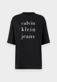Černé bavlněné tričko s oversize střihem, které na sobě má vertikální bílé texty: "calvin klein jeans" v tučném, moderním písmu. Krátké rukávy a kulatý výstřih.