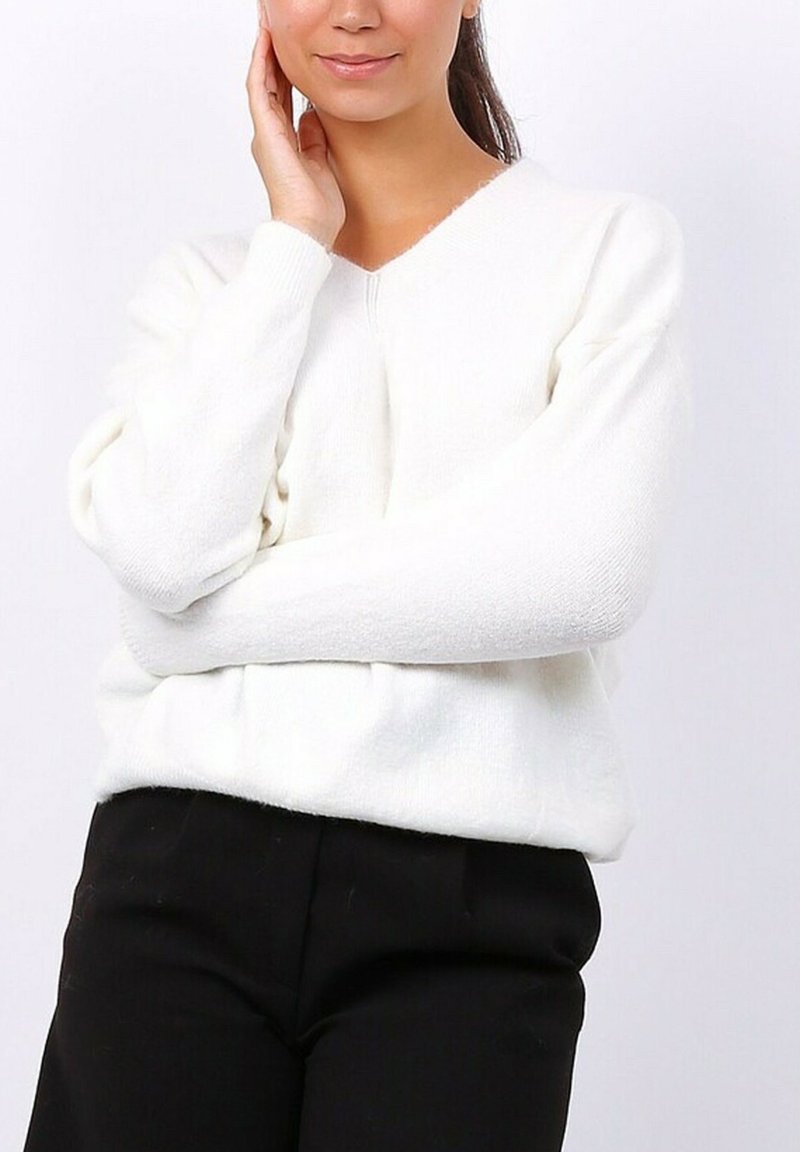 Femme portant un pull à manches longues blanc à col en V et un pantalon noir, avec un bras croisé et l'autre main touchant son visage.