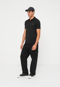 Čierne polo tričko so zlatým logom ako akcent, kombinované s čiernymi cargo nohavicami s bočnými vreckami a čiernymi teniskami s bielou podrážkou.