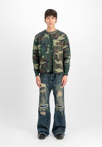 Giovane uomo con giacca mimetica verde, jeans larghi e strappati con toppe, e scarpe nere, in piedi davanti a uno sfondo bianco.
