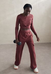 Burgundi hosszúujjú cropped top, csavart részlettel, laza burgundi nadrággal párosítva. Az illető egy fekete kézisúlyt tart, világos sportcipőt viselve.