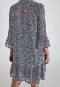 Vestido de estampado floral en tejido azul translúcido con mangas tres cuartos y dobladillo con volantes. Presenta patrones florales de color crema en todo el vestido.