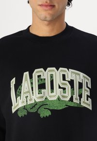 Sudadera de algodón negra con un gran gráfico en blanco y verde de "LACOSTE", con un diseño texturizado de cocodrilo verde detrás del texto.