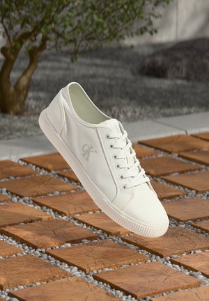 Baskets blanches Calvin Klein suspendues au-dessus d'un bloc en bois et d'un sol extérieur en gravier, avec un arrière-plan d'arbre et de béton.