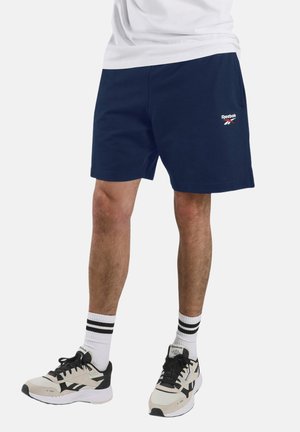 Uomini che indossano pantaloncini Reebok blu navy, calze bianche con strisce nere e scarpe da ginnastica Reebok bianche e nere, in piedi contro uno sfondo chiaro.