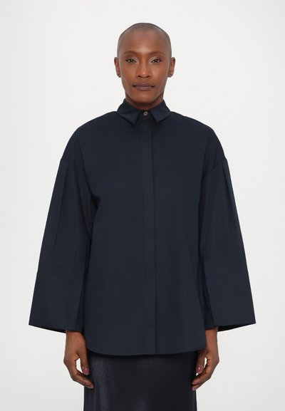 Ναυτικό μπλε oversized πουκάμισο με γιακά, κούμπωμα με κουμπιά και φαρδιές μανσέτες. Λείο ύφασμα με σταθερή γραμμή, χωρίς ορατά σχέδια.