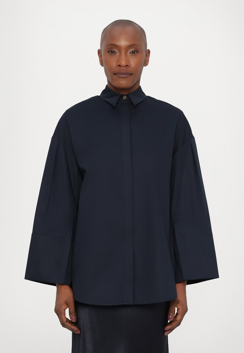 Camicia oversize blu navy con colletto, chiusura a bottoni e maniche ampie. Tessuto liscio con una forma strutturata, senza motivi visibili.