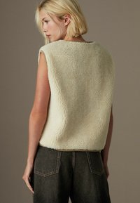 Creme farvet falsk pels ærmeløs top med en blød tekstur, rund halsudskæring og sænket armhuller, parret med mørke denim high-waisted bukser.