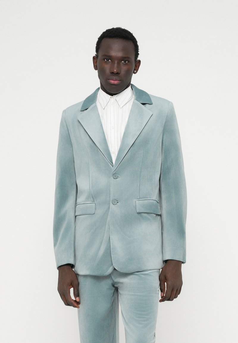 Svijetloplavi baršunasti odijelo s reverima, dva gumba i dva prednja džepa. Nosit će se preko bijele prugaste košulje, s glatkom teksturom u cijelosti.