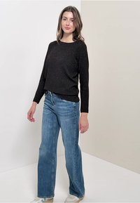 Czarny, błyszczący sweter z długim rękawem w połączeniu z jasnoniebieskimi spodniami o szerokich nogawkach i białymi trampkami, stojący na neutralnym tle.