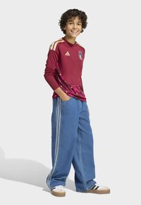 Maroon long-sleeve jersey with gold accents and logo, paired with wide blue denim pants featuring side stripes and sneakers.