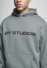 Felpa grigia realizzata in morbido tessuto, con tasca frontale e logo stampato "2Y STUDIOS" in grandi lettere nere sulla parte anteriore e posteriore.