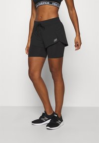 Kvinna som bär en svart New Balance sport-bh, lager av svarta träningsshorts och svarta löparskor, står mot en vit bakgrund.