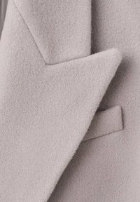 Lysegråt uldstof med en struktureret overflade, med et markant reversdesign og en subtil lomme-detalje, der er synlig på siden.