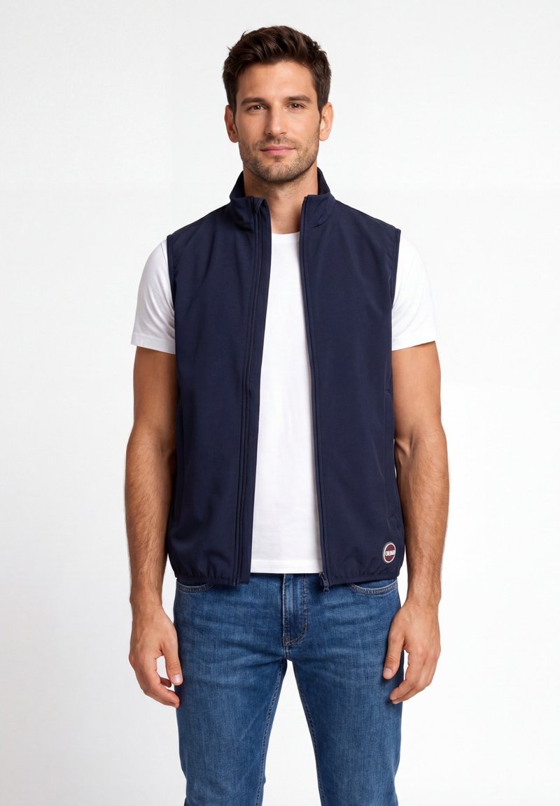 Egy férfi, aki tengerészkék ujjatlan mellényt visel fehér póló felett és kék farmert, egy egyszínű fehér háttér előtt áll.