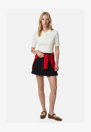 Kvinde iført hvid kortærmet bluse med krave, sort plisseret nederdel, rød sweater bundet om livet og brune Mary Jane-sko, stående på hvid baggrund.