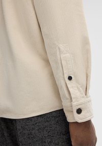 Light beige corduroy shirt with a ribbed texture, featuring buttoned cuffs and black buttons, extending to a plain hem at the bottom.
