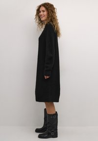 Juoda, perDidelė megzta suknelė su ilgomis rankovėmis ir banguotais kraštais. derinama su juodais, iki kelių siekiančiais botais, kurie turi sagties akcentus.