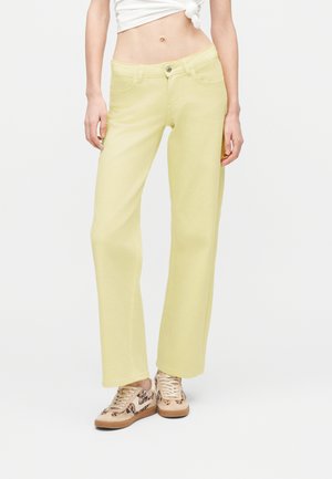 Žena v kalhotách s vysokým pasem světle žluté barvy a širokými nohavicemi, obutá v béžových teniskách se zvířecím vzorem, stojící před jednobarevným bílým pozadím.