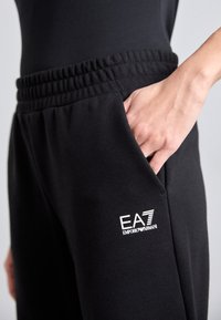 Melni sporta bikses ar elastīgu jostas vietu, sānu kabatām un baltu EA7 Emporio Armani logo uz kreisā augšstilba.