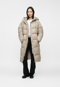 Beige puffer coat with a hood, featuring a quilted design, front zipper closure, and two side pockets. Worn over black trousers and shoes.