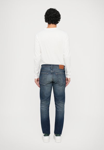 Modré denim jeans so strihom rovného strihu, s zadnými vreckami a pieskovou koženou nášivkou. Nosené s bielou košeľou s dlhým rukávom a bielymi teniskami.