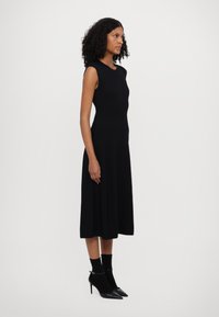 Vestido preto com bainhas, com corpete justo e saia rodada, usado com meias pretas até o tornozelo e sapatos de salto agudo. Textura suave e design elegante.