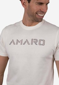 Camiseta de color crema con mangas cortas y cuello redondo, con letras bordadas en azul texturizado que dicen "AMARO". Tela suave.