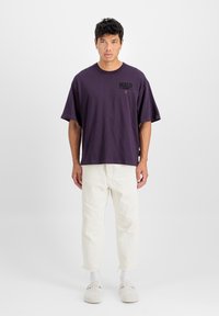 T-shirt en coton violet avec un col ras du cou, des manches courtes et un design graphique. Associé à un pantalon de couleur claire et des chaussures grises.