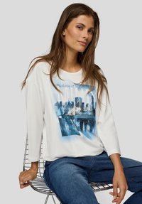 Junge Frau mit langen braunen Haaren, trägt ein weißes langärmeliges Shirt mit Grafik und blaue Jeans, sitzt auf einem Metall-Drahtstuhl vor einem schlichten Hintergrund.