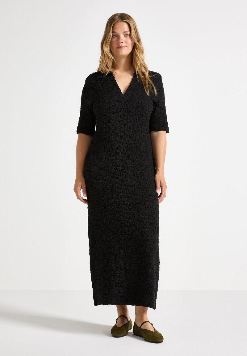 Robe noire avec une finition texturée, décolleté en V et manches courtes. Coupe mi-longue accompagnée de chaussures plates vert foncé. Design simple et forme épurée.