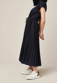 Tummansininen pleatattu midimekko kevyestä kankaasta, jossa on narukiristys vyötäröllä, yhdistettynä valkoisiin lenkkareihin, joissa on monivärisiä yksityiskohtia.