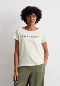 T-shirt à manches courtes de couleur crème clair avec le texte noir imprimé "FAITES UNE DIFFÉRENCE." Tissu en coton doux et lisse avec une coupe décontractée.