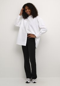 Chemise blanche à manches longues avec col à boutons et poche poitrine, associée à un pantalon noir ajusté et des chaussures de sport grises. Fond uni.