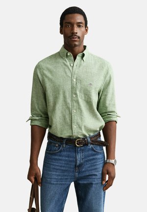 Uomo che indossa una camicia verde chiaro con bottoni e maniche arrotolate, jeans blu, cintura marrone intrecciata, orologio argentato e tiene una tracolla marrone.