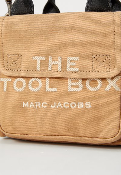 Geantă din pânză cu un ton bej, având accente din piele neagră, cu text brodat alb: "THE TOOL BOX" și "MARC JACOBS." Cusăturile sunt vizibile.