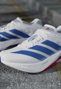 Λευκά και μπλε παπούτσια τρεξίματος Adidas Adizero με κόκκινες λεπτομέρειες που ακουμπούν σε μια υφή εξωτερικής επιφάνειας.