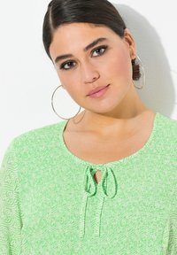 Blouse verte texturée avec un col rond et un détail à nouer. Manches longues avec une insert en mesh à motif.